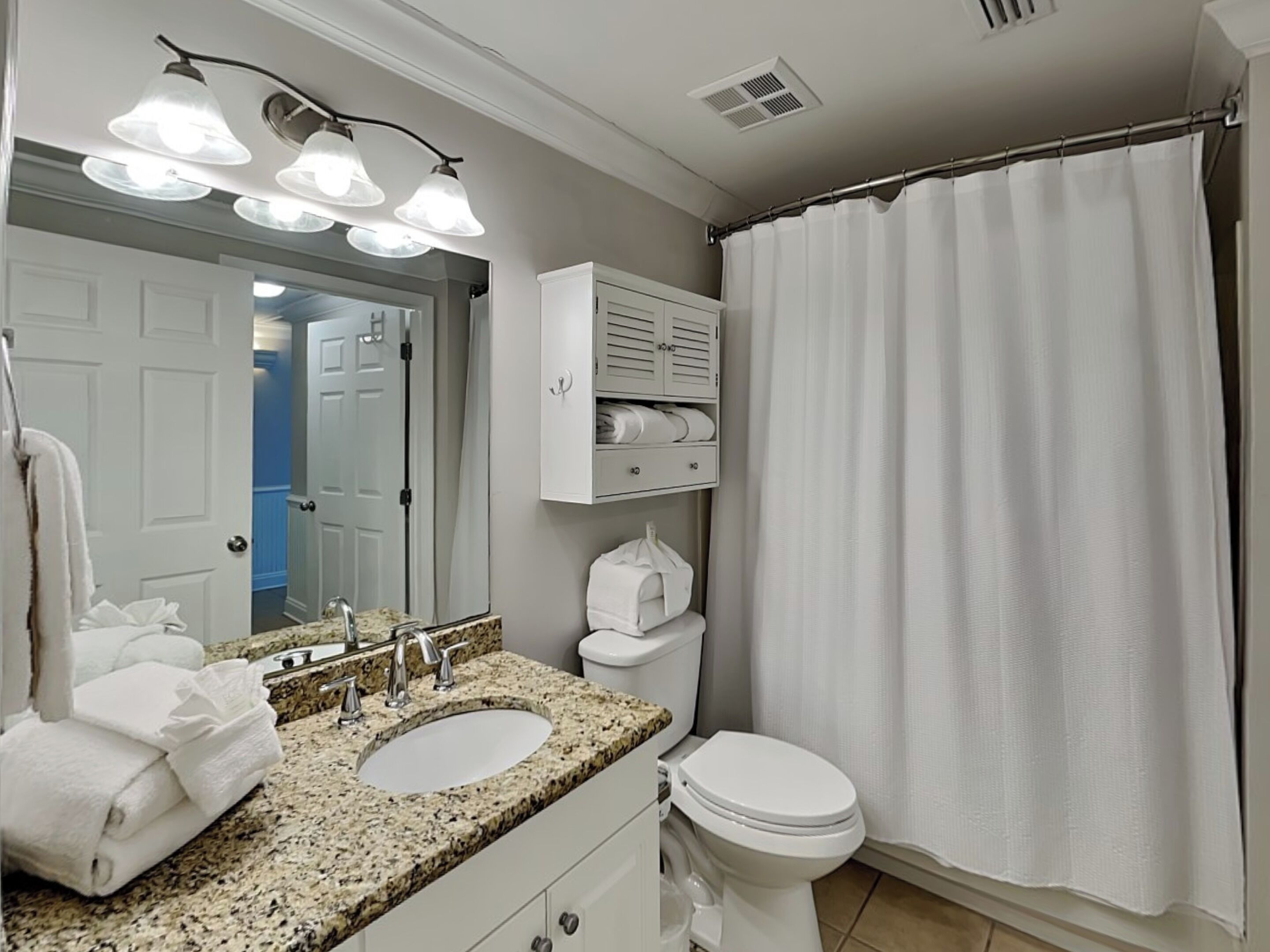 A bathroom with a granite countertop sink, a white toilet, a shower curtain, and a mirror with three lights.