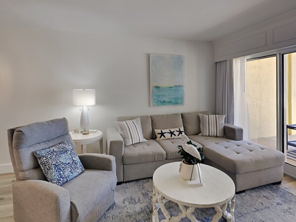 Un salon moderne avec un ensemble de canapés gris, une table basse ronde et une table d’appoint avec une lampe. Un tableau orne le mur et une grande fenêtre offre une vue sur l’extérieur.