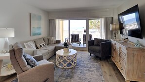 A spacious living room with a large sofa, a smaller armchair, a coffee table, and a TV mounted on the wall. - Palm View | Serene Views, Cozy Interior, Near Area Fun! (Gulf Shores)