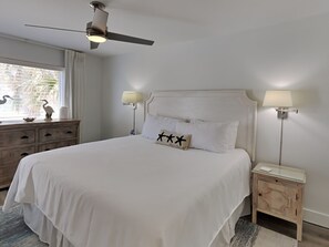 Une chambre à coucher comprenant un lit, une table de chevet, une lampe, un ventilateur de plafond et une fenêtre avec des rideaux.