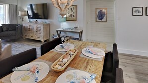 A dining area with a wooden table set for four, surrounded by dark chairs. A chandelier hangs above the table, and a television is mounted on the wall to the left. - Palm View | Serene Views, Cozy Interior, Near Area Fun! (Gulf Shores)
