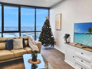 Ein modernes Wohnzimmer mit einer Ledersofa, einem Glastisch, einem Weihnachtsbaum, einem Flachbildfernseher und Blick auf die Stadt durch große Fenster.
