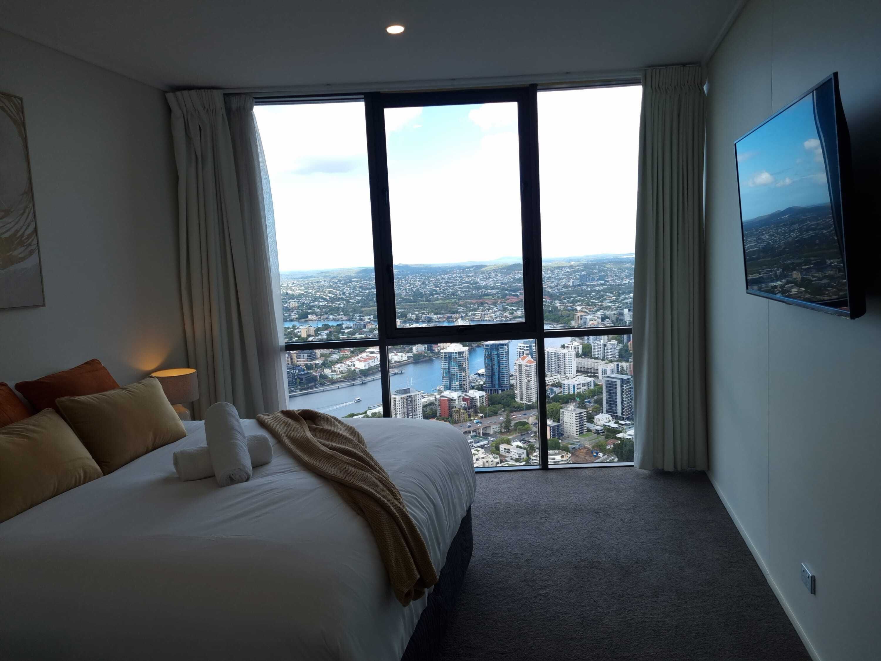 A hotel room with a large window offering a city view, a bed with pillows, and a flat-screen TV mounted on the wall.