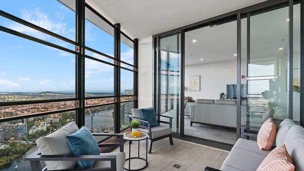 A modern apartment with large windows offering a city view, a balcony with seating, and a kitchen area visible through glass doors. - Luxury 4 Bedroom Sub Penthouse with Ocean Views (Brisbane City)