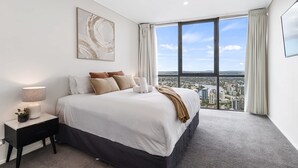 A modern hotel room with a large bed, a nightstand, a lamp, and a city view through a large window. - Luxury 4 Bedroom Sub Penthouse with Ocean Views (Brisbane City)