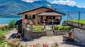 Una casa de fachada de piedra, un patio con mobiliario exterior y vistas a un lago y montañas.