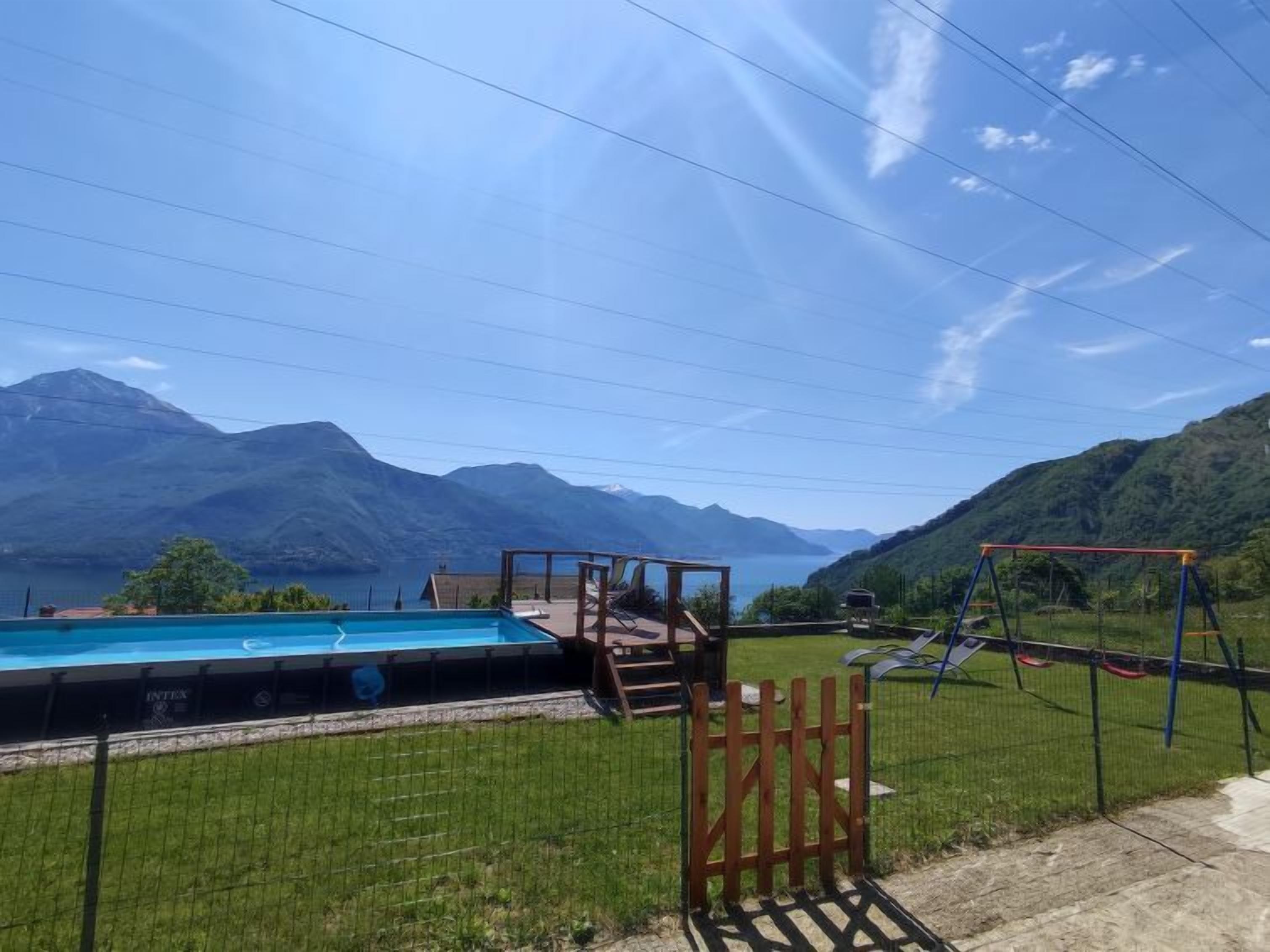 A swimming pool with a wooden deck and a swing set, surrounded by a grassy area and mountains in the background.