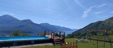 A swimming pool with a wooden deck and a swing set, surrounded by a grassy area and mountains in the background.