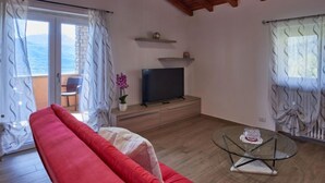 Una sala de estar con un sofá rojo, una mesa de centro de vidrio, un televisor sobre un mueble de madera y un balcón con vista al mar.