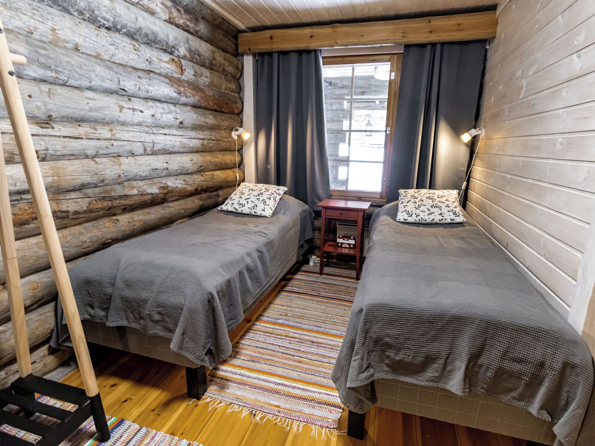Une chambre aménagée dans un chalet en rondins, avec deux lits, une échelle en bois, une petite table et une fenêtre avec des rideaux.