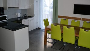 A compact kitchen with white cabinets, a microwave, a coffee maker, and a sink. There is a dining area with a wooden table and green chairs. A flat-screen TV is mounted on the wall.