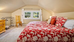 Ein Schlafzimmer mit schrägverdachter Decke, einem Bett mit roter Blumenbettwäsche, einem Holzstuhl, einer Holzkommode und einem Fenster mit weißen Vorhängen.