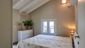 Een slaapkamer met een bed, een dressoir, een kamerplant, een raam en een wandlamp.