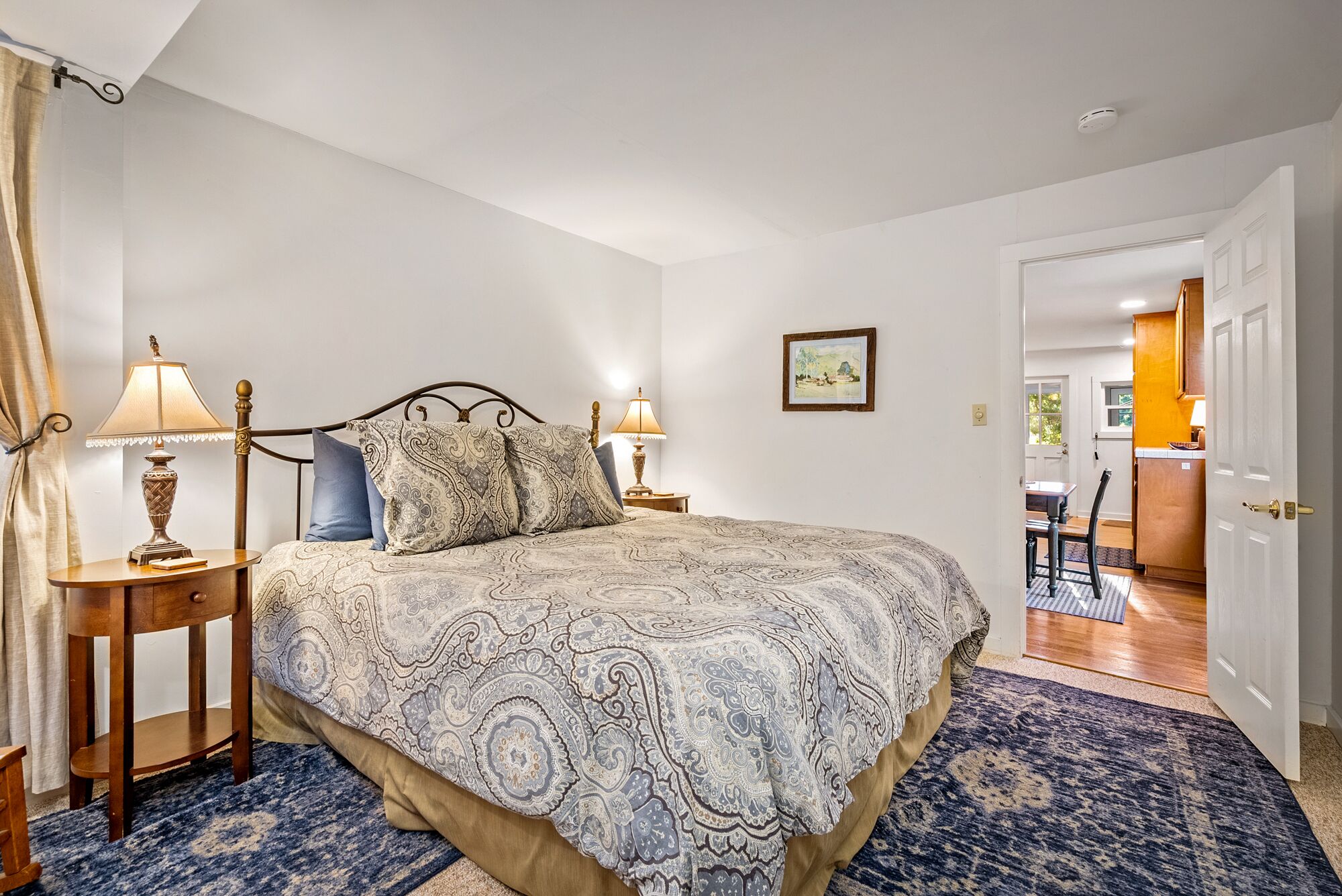 A bedroom with a large bed, two bedside tables with lamps, a blue patterned rug, and a door leading to a dining area.