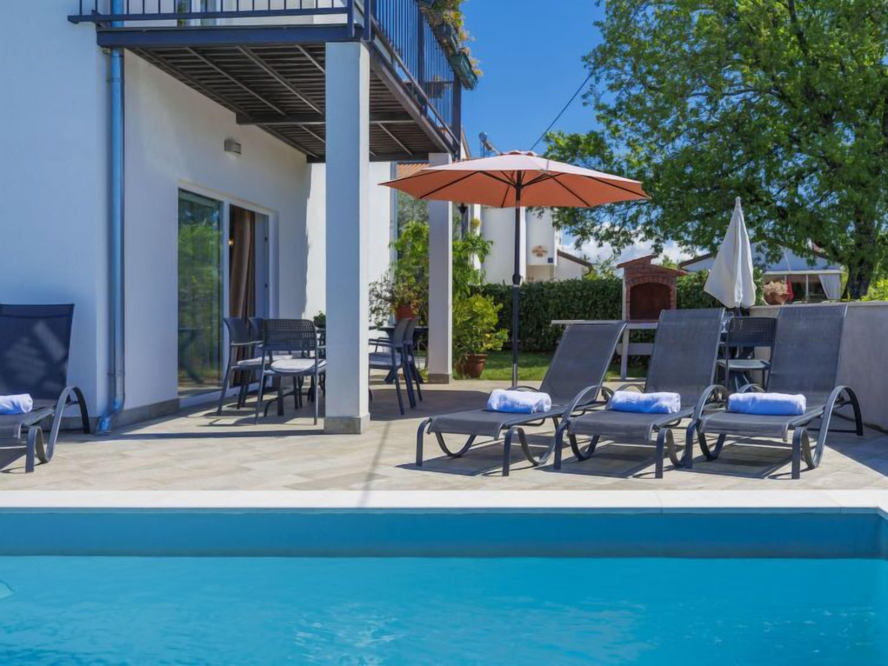 A poolside area with lounge chairs and a table, a covered patio, and a clear blue pool.