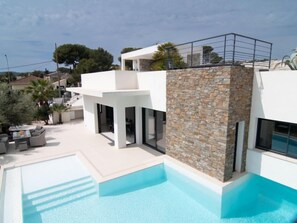 A modern two-story house with a swimming pool, stone wall, and outdoor seating area.