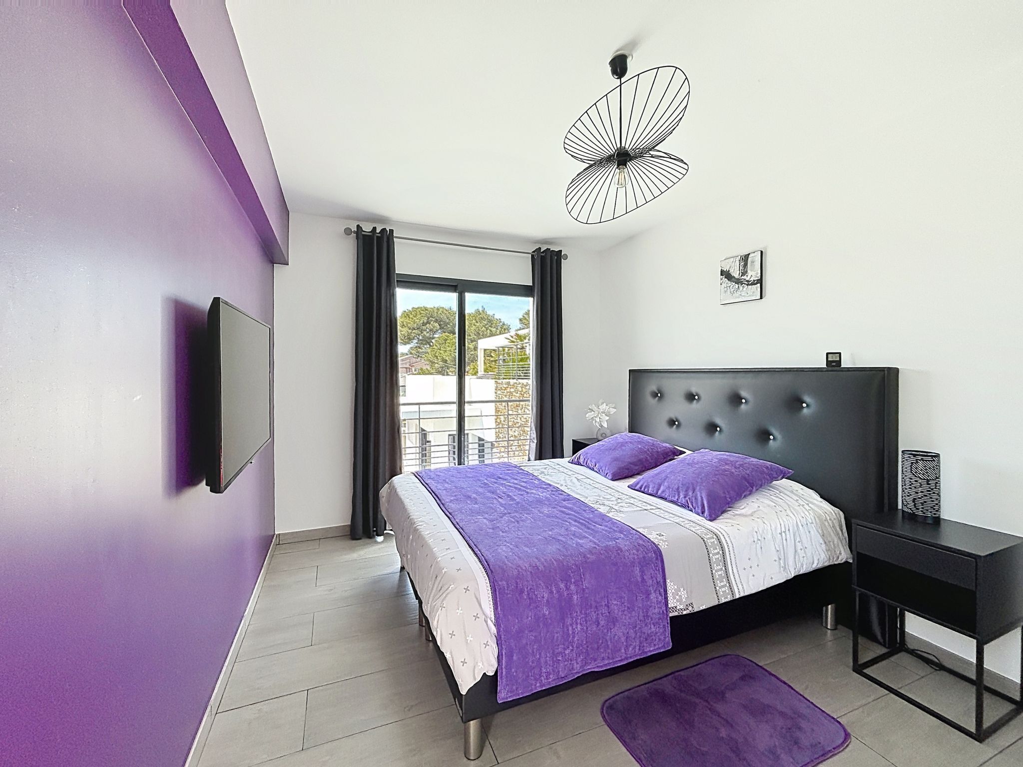 Un dormitorio moderno con cama, una mesita de noche negra, televisor montado en la pared y una ventana con cortinas.