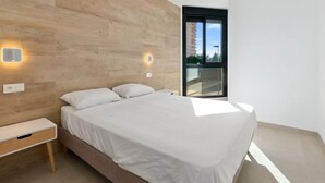 Une chambre moderne avec une tête de lit en bois, un grand lit avec une literie blanche, une table de chevet et une fenêtre donnant sur des immeubles.