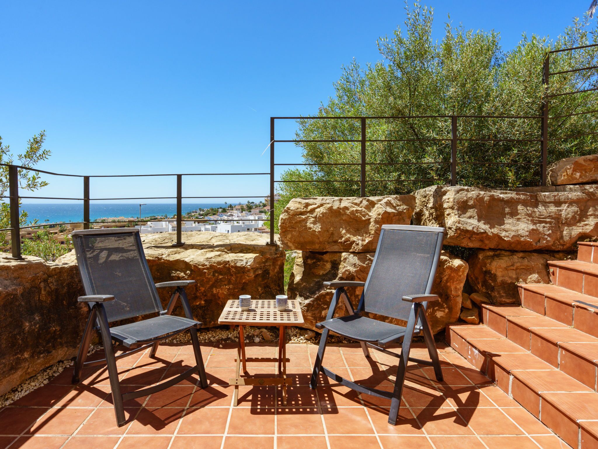 Eine Terrasse mit zwei Stühlen und einem Tisch, mit Blick auf eine Küstenstadt und das Meer.