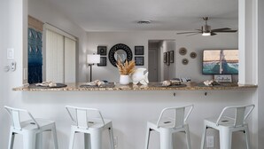 A kitchen with a granite countertop, white bar stools, a ceiling fan, and a TV displaying a desert landscape. - 5027 Fairfield Dr - Paradise Palms 3 Bedroom Home by RedAwning (Fort Myers)