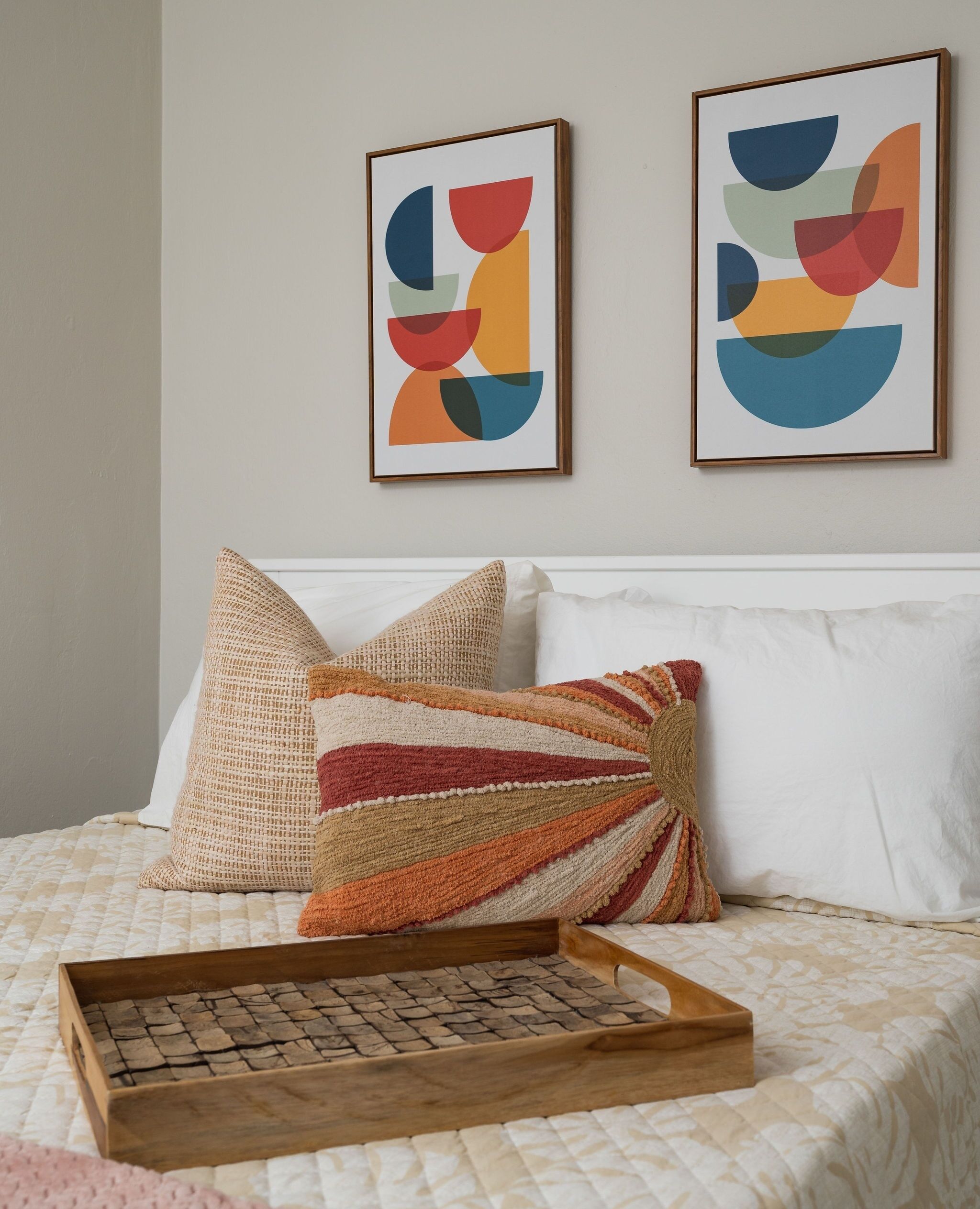 A neatly made bed with a wooden tray, two pillows, and two abstract paintings on the wall.