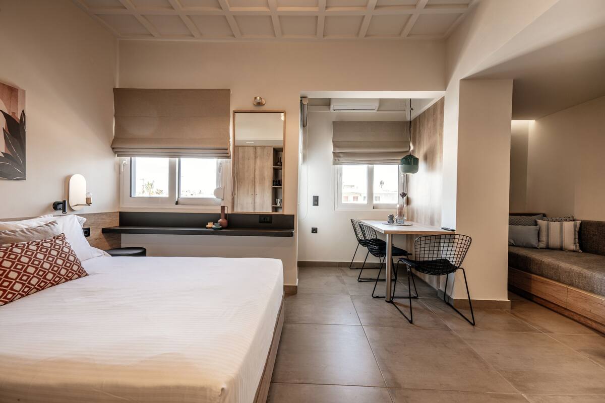 Una habitación de hotel moderna con una cama grande, un escritorio con una silla, un sofá y vistas a la ciudad desde las ventanas.