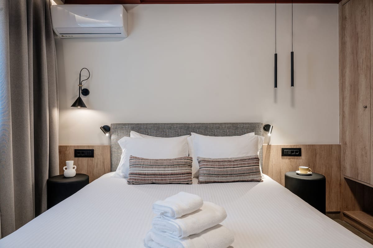 Una cama bien hecha con sábanas blancas, dos almohadas y una pila de toallas dobladas. Hay una mesita de noche con una taza y un pequeño florero. La habitación tiene un diseño moderno con paneles de madera y aire acondicionado instalado en el techo.