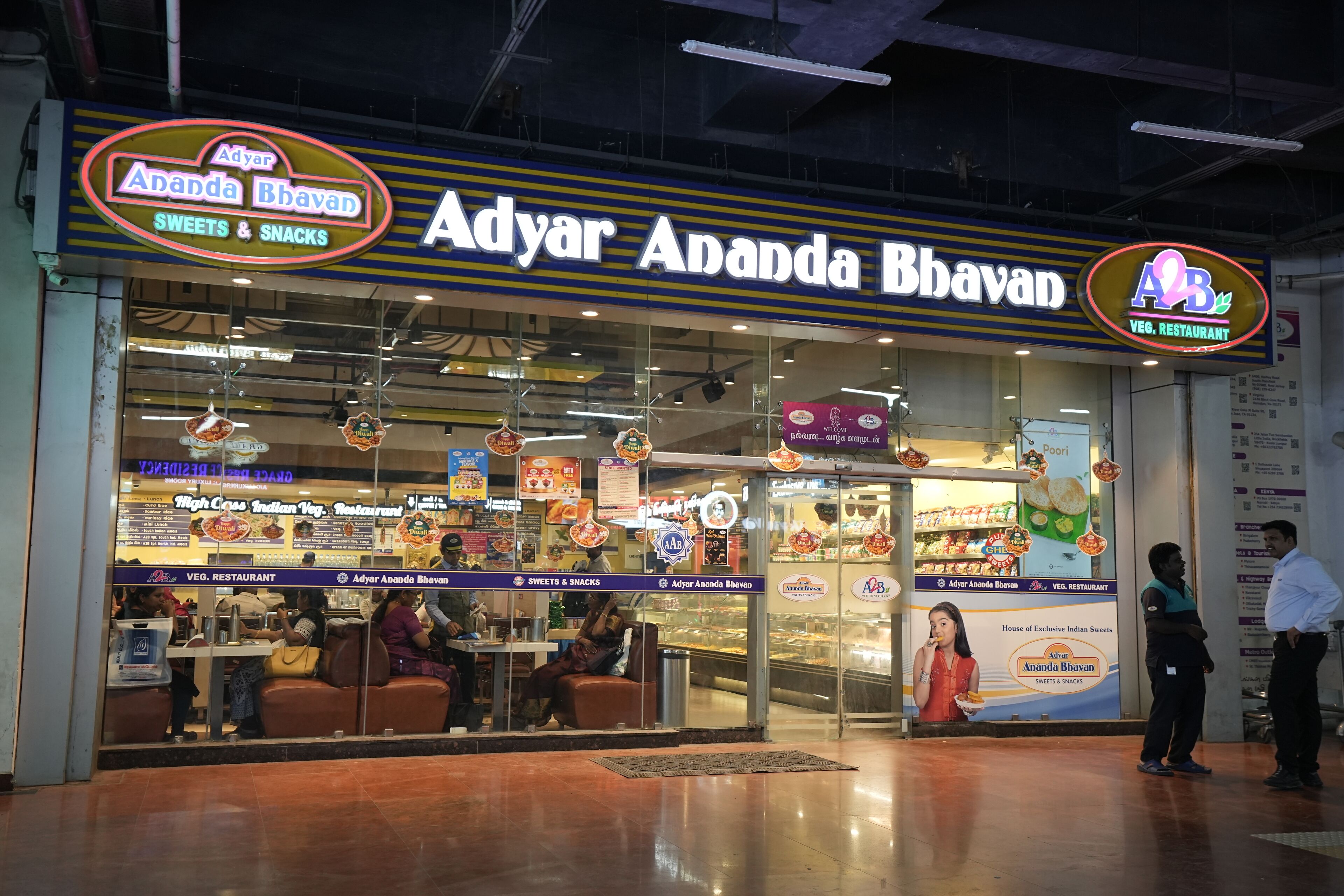 A restaurant named Adyar Ananda Bhavan with a signboard and a glass display of sweets and snacks.