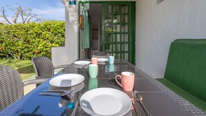 A dining table set with plates, cups, and cutlery, surrounded by chairs, in a covered outdoor area with a green door and a hedge in the background.