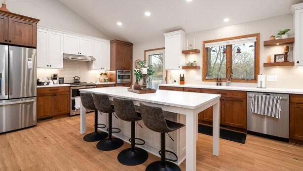 A modern kitchen with wooden cabinets, stainless steel appliances, a central island with bar stools, and a window with a view of trees.