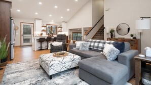 A spacious living room with a grey sectional sofa, a patterned ottoman, and a large rug. A kitchen with white cabinets and a dining area with bar stools is visible in the background.