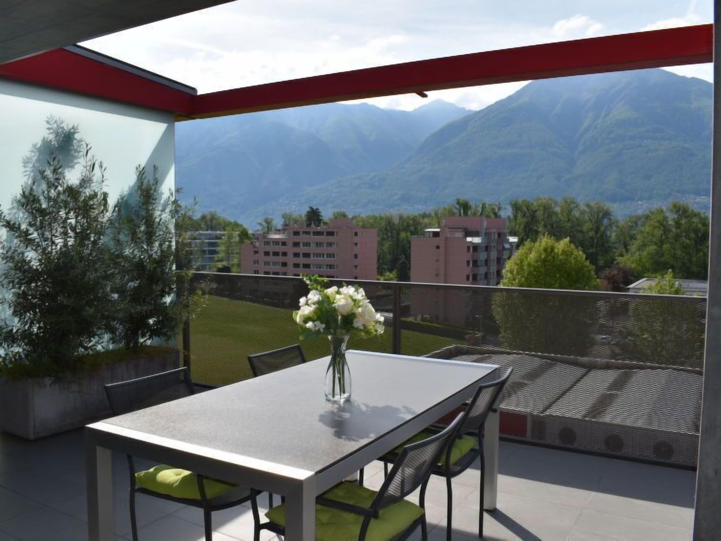 A balcony with a table and chairs, a vase with flowers, and a view of mountains and buildings.