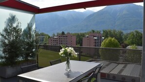 Un balcone con tavolo e sedie, un vaso con fiori e vista sulle montagne e sugli edifici.
