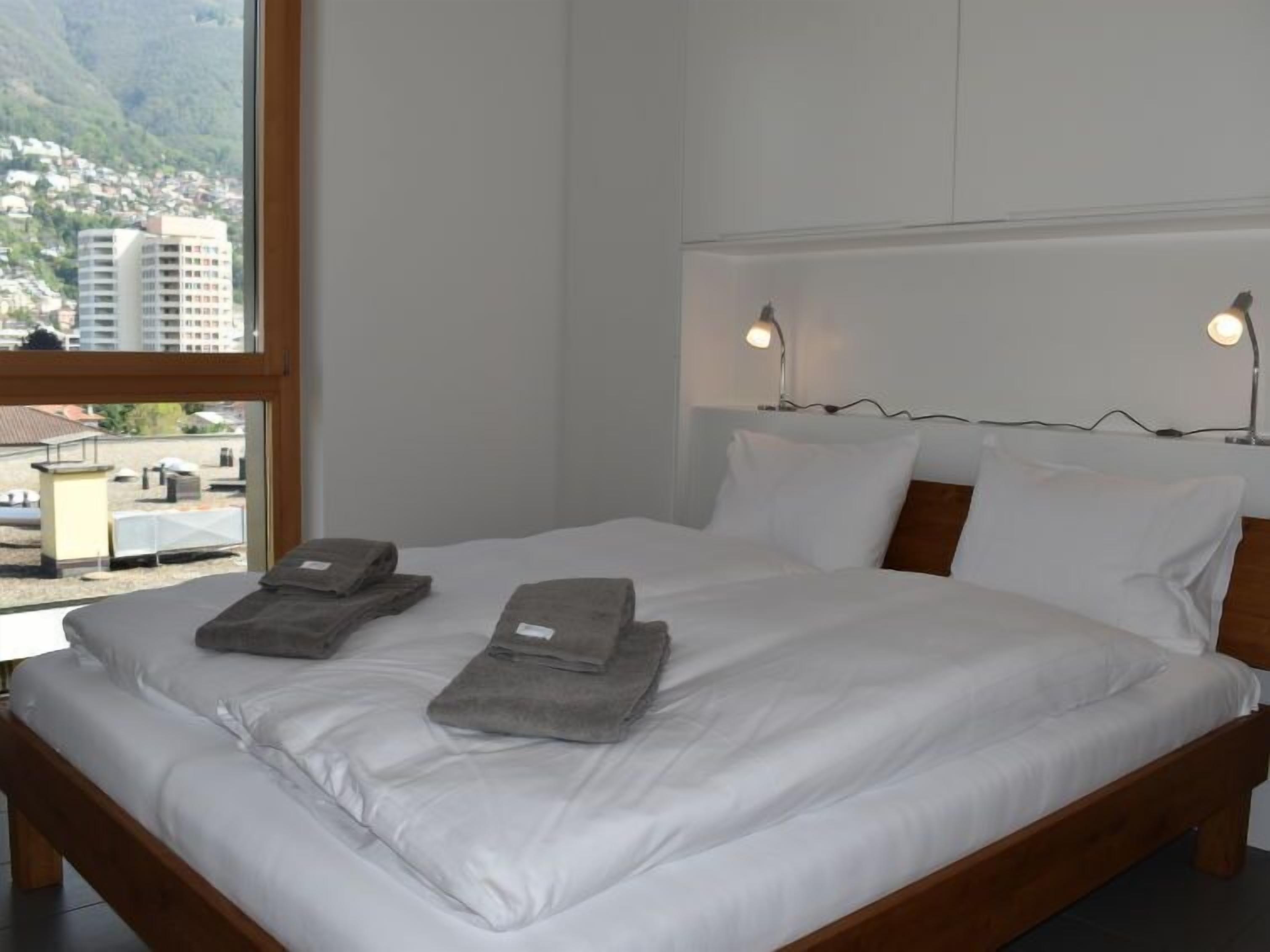 A neatly made bed with white linens and two folded towels, a wooden headboard, and two bedside lamps.