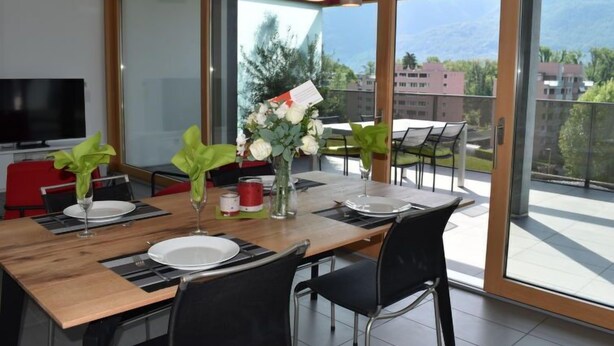 Zona pranzo moderna con tavolo in legno, sedie nere e una grande finestra con vista sulle montagne.