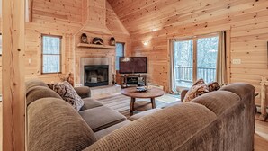 Interior acogedor de una cabaña de madera con chimenea, televisor y un ventanal grande con vistas a un paisaje nevado.