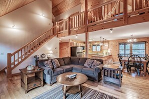 A spacious living area with a wooden staircase, a sectional sofa, a dining table with chairs, and a kitchen in the background.