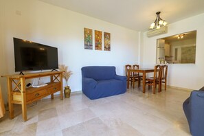 A living room with a flat-screen TV, a blue sofa, a wooden dining table with chairs, and a small vase with dried plants.