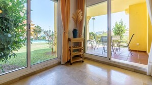 A room with large sliding glass doors leading to a balcony, a small wooden shelf with a vase, and a view of a pool and greenery outside. - Elegant Mijas Costa Holiday Home for 4 (Mijas Costa)