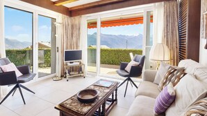 Amplia sala de estar con sofá, sillones, mesa de centro y televisor sobre un mueble. La habitación cuenta con grandes puertas corredizas de vidrio que dan a un balcón con vista a la montaña.
