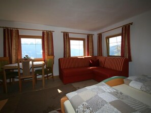 Quarto com cama, sofá vermelho, cadeira de madeira, mesa e janelas com cortinas.