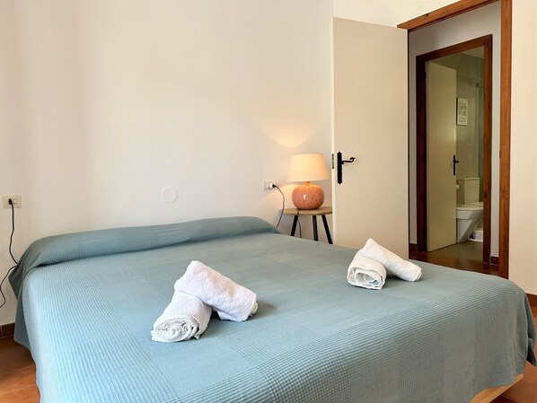 A neatly made bed with two rolled towels, a bedside lamp, and a door leading to a bathroom.