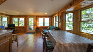 A cabin interior with a dining table, chairs, a TV, and large windows offering a view of the outdoors.