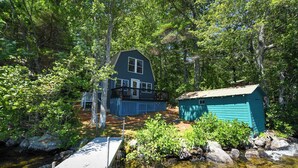 A cabin with a dock extending into a clear stream, surrounded by dense forest.