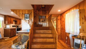 A wooden interior with a staircase leading to a loft bedroom, a kitchen area, and a living space with a wicker chair.