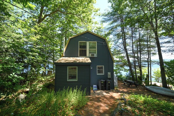 A small, dark green cabin with a single window and a door, surrounded by dense trees and a dirt path leading to a picnic table.