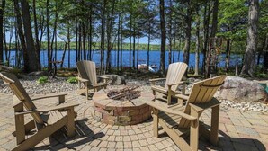 Een houten terras met Adirondack-stoelen en een vuurkuil, met uitzicht op een meer met boten.