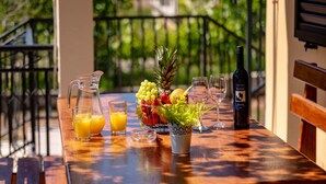 Ein Holztisch mit Obstschale, einer Weinflasche, Gläsern und einem Orangensaftkrug auf einem Balkon mit Geländer und Grünpflanzen im Hintergrund.