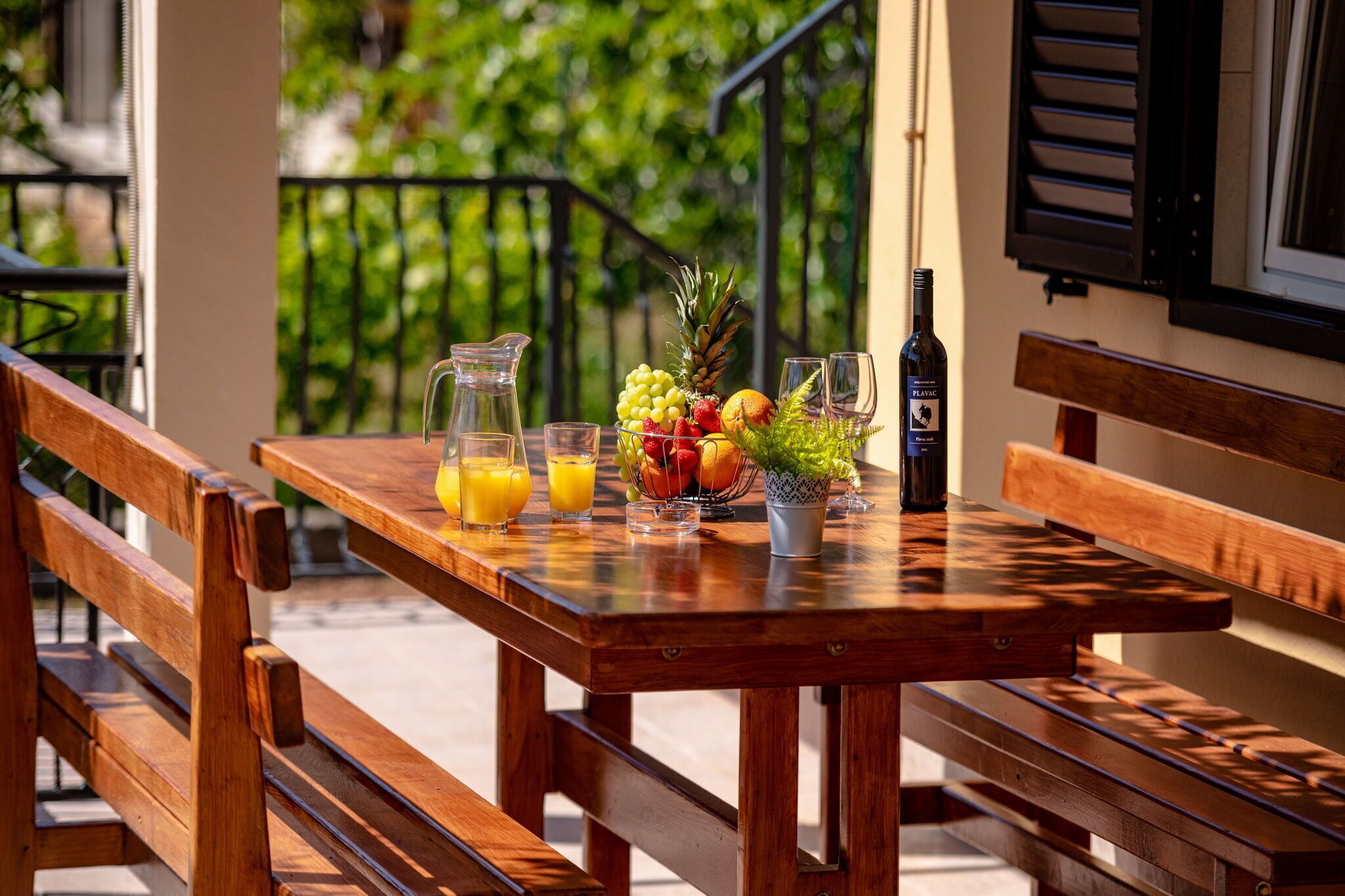 A wooden table with a pitcher of orange juice, glasses, a bottle of wine, and a bowl of fruit on a balcony.