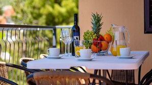 Une table dressée avec une bouteille de vin, des verres, un panier de fruits et un pichet de jus d’orange, sur un balcon d’où l’on aperçoit des arbres.